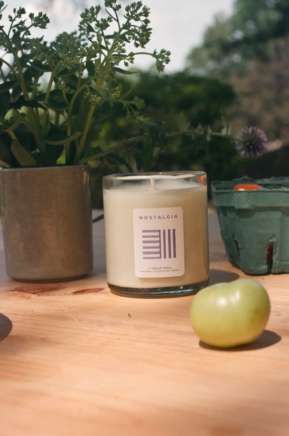 Candle with a label on a wooden surface next to a plant and a green fruit