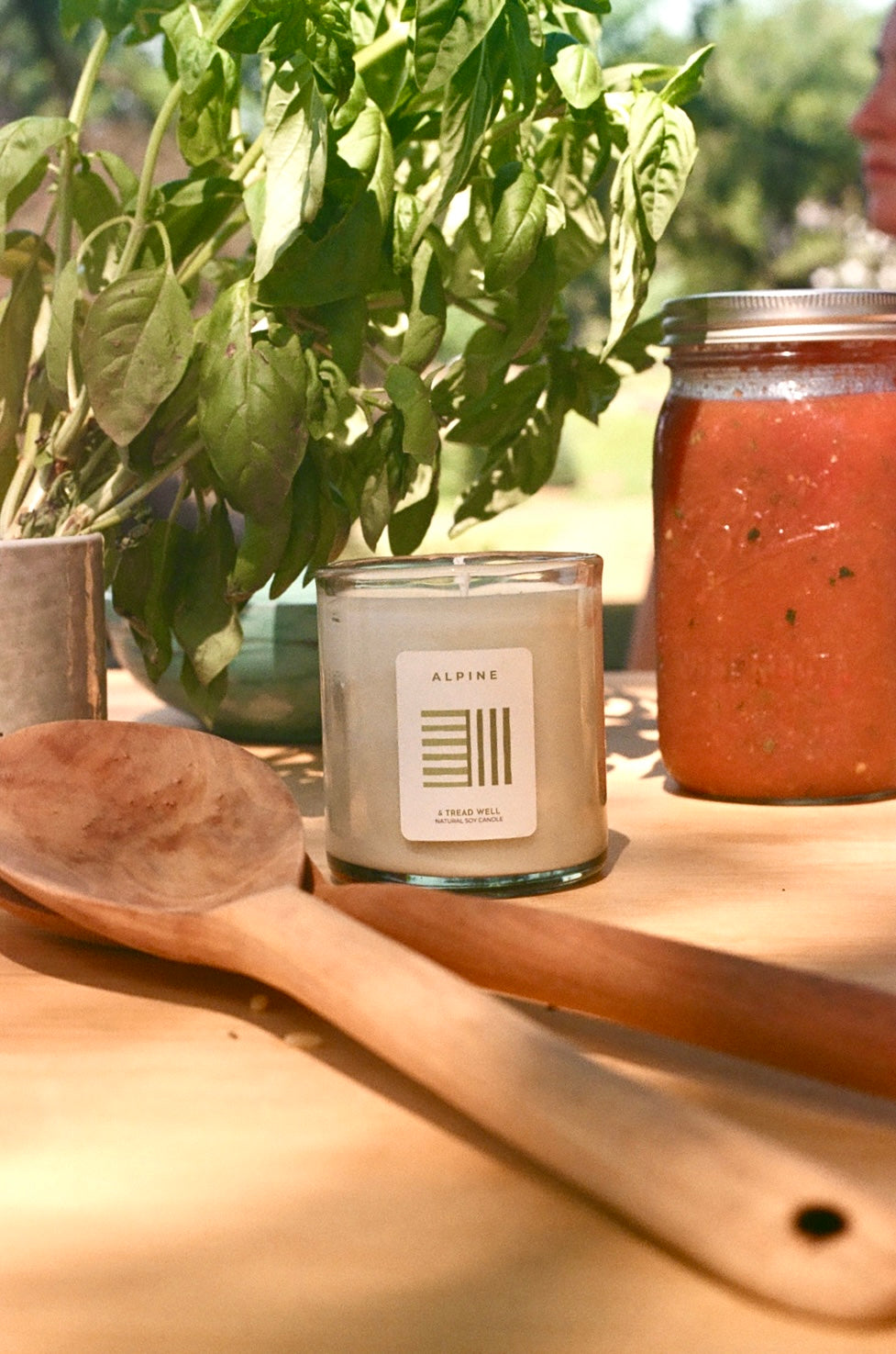 Candle with a label, wooden spoon, and jar of red sauce on a wooden surface with plants in the background