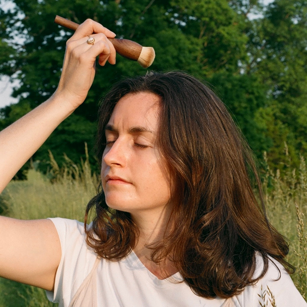 Woman in a field holding a stick and a small container, surrounded by greenery.