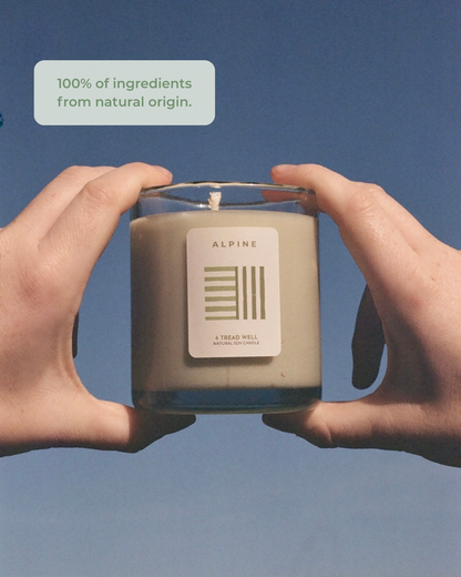 Two hands holding a candle labeled 'Alpine' against a blue background with text about natural ingredients.