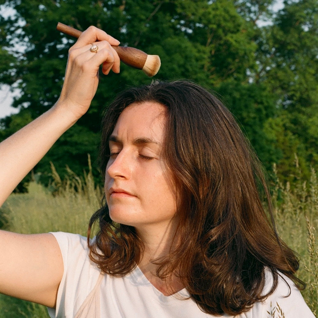 Woman in a field holding a stick and a small container, surrounded by greenery.