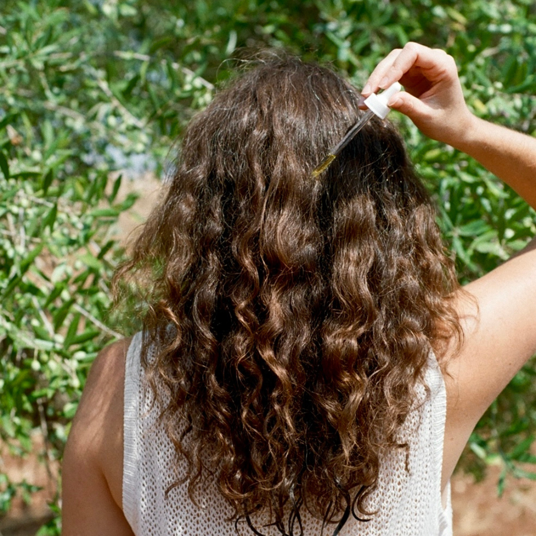 Person applying hair product to their curly hair outdoors with greenery in the background