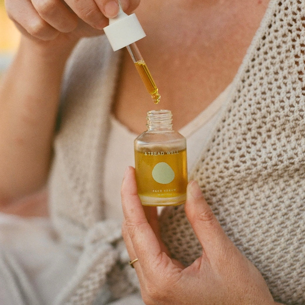 A close-up lifestyle shot of a woman holding a small glass bottle of golden-yellow serum and using a dropper to apply it. She is wearing a beige knit cardigan and a cream tank top, with a soft-focus outdoor background.
