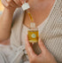 A close-up lifestyle shot of a woman holding a small glass bottle of golden-yellow serum and using a dropper to apply it. She is wearing a beige knit cardigan and a cream tank top, with a soft-focus outdoor background.