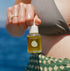 A person outdoors by the ocean holds a small bottle of & TREAD WELL Face Serum. The bottle contains a golden-yellow serum and has a white dropper cap.