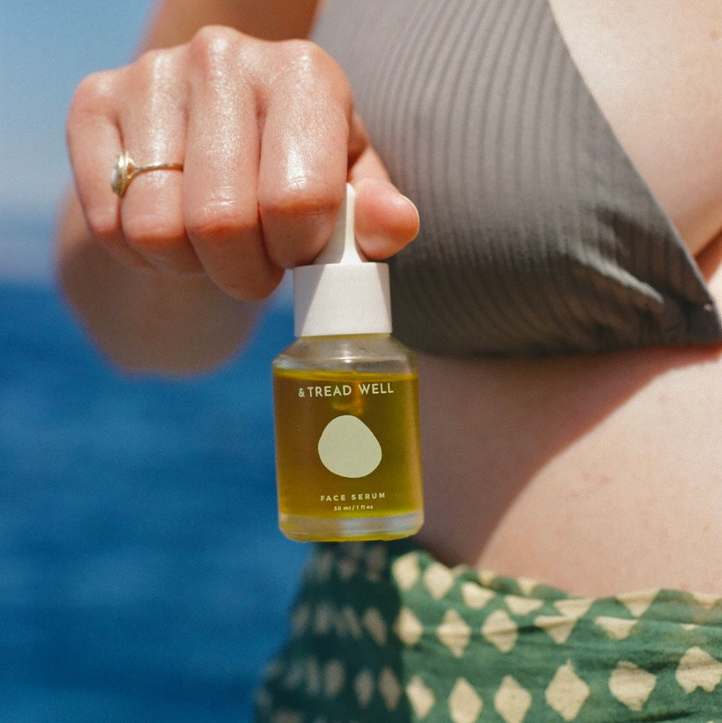 A person outdoors by the ocean holds a small bottle of & TREAD WELL Face Serum. The bottle contains a golden-yellow serum and has a white dropper cap.