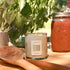 Candle with a label, wooden spoon, and jar of red sauce on a wooden surface with plants in the background