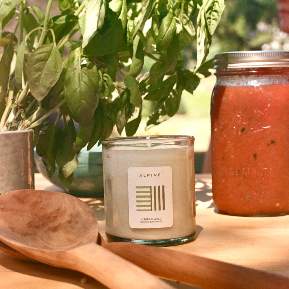 Candle with a label, wooden spoon, and jar of red sauce on a wooden surface with plants in the background