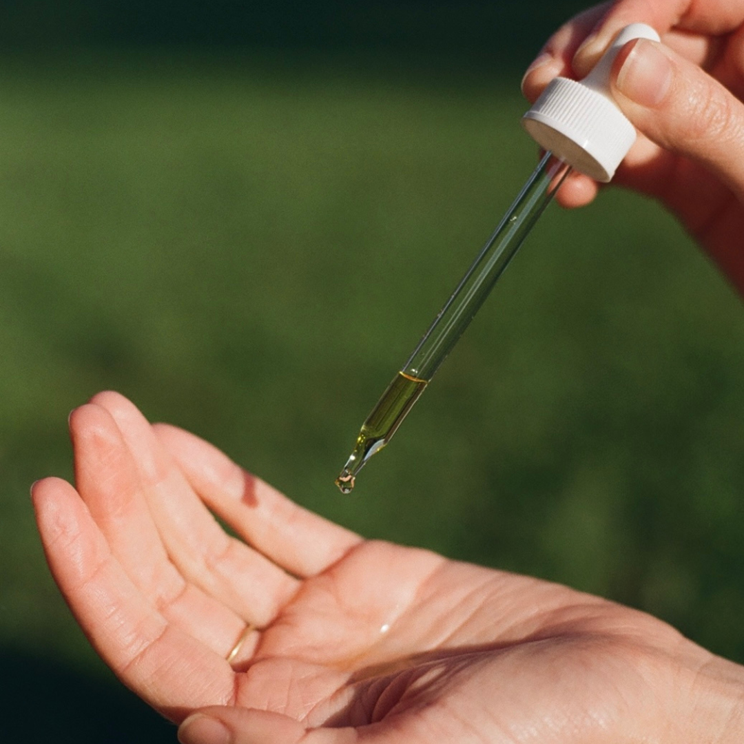 Hand holding a dropper with a drop of liquid over another hand against a blurred green background