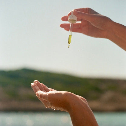Hand holding a dropper with liquid over another hand by a body of water