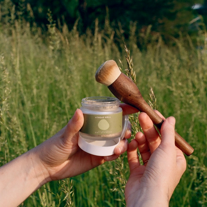 Person holding a jar of powder and a wooden applicator against a natural background